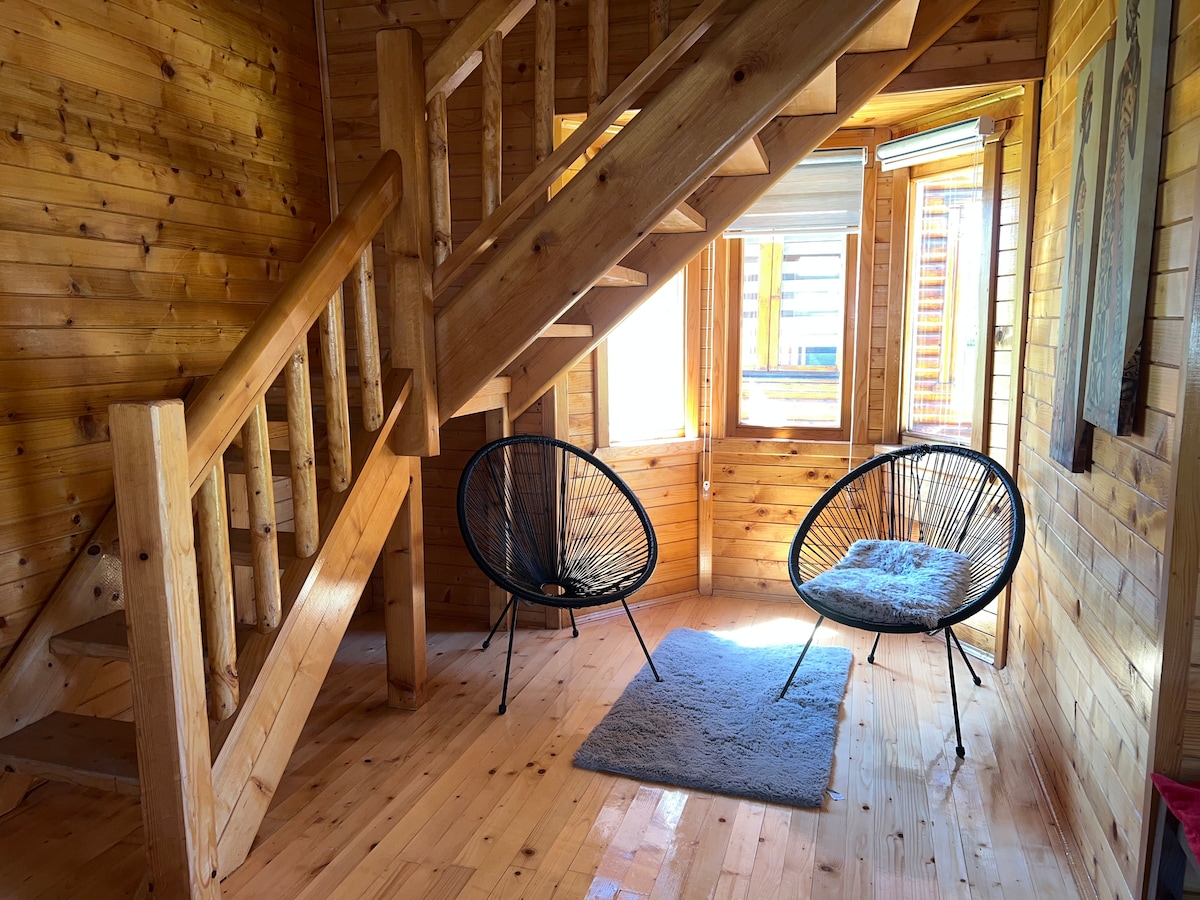 A cozy corner is created by two black acapulco chairs placed on a soft rug. Light enters through large windows, illuminating the wooden walls and staircase, enhancing the natural warmth of the space.