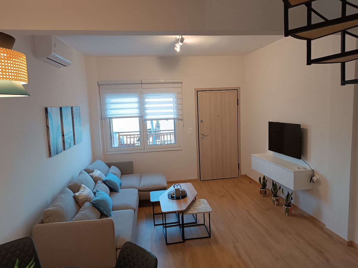 A bright living area is furnished with a light gray sectional sofa, accented by blue cushions. A wooden coffee table is placed in front, with decorative items on it. A wall-mounted television can be seen opposite the sofa, and large windows allow natural light to enter the space.