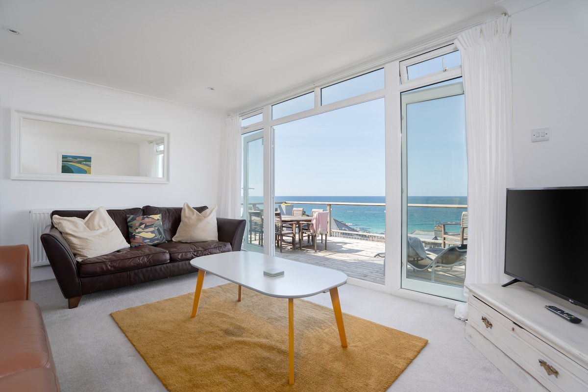 A bright lounge space features large glass doors that open to a stunning seaside view. Comfortable seating is arranged around a central coffee table, with a plush area rug underneath. Natural light fills the room, creating an inviting atmosphere enhanced by soft white curtains.