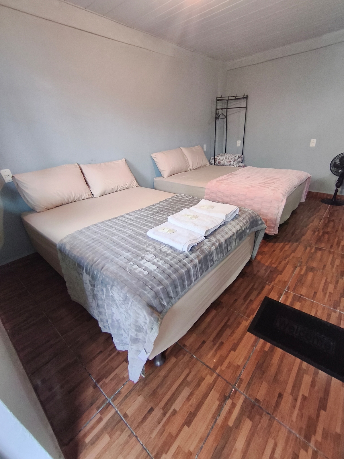 The sleeping area contains two spacious beds, one with a light-colored blanket and the other with a pink quilt. Fresh towels are neatly folded on top. A fan is positioned nearby, and a clothing rack is available for storage against the wall.