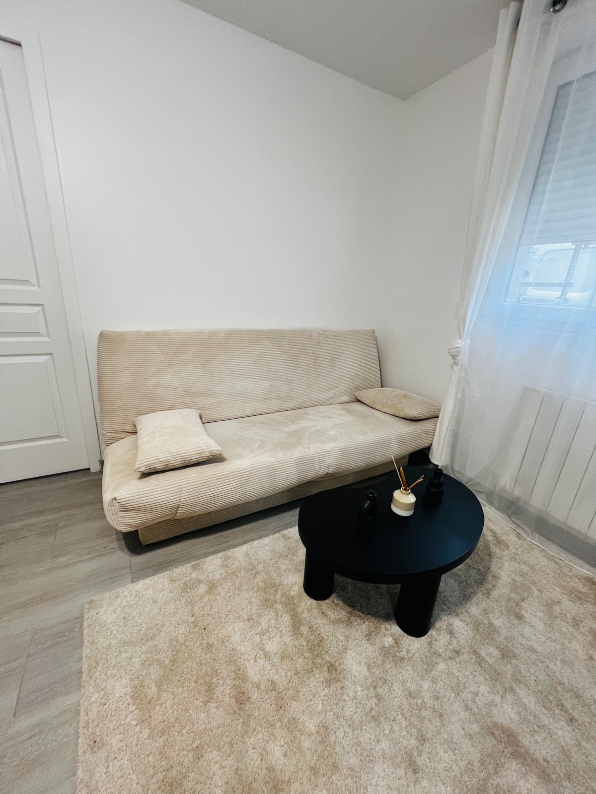 A light-filled corner of the living space is shown, featuring a soft beige sofa with a small accent pillow. A round black table sits in front, accompanied by a decorative item. A soft area rug complements the space, while a window allows natural light to filter in.