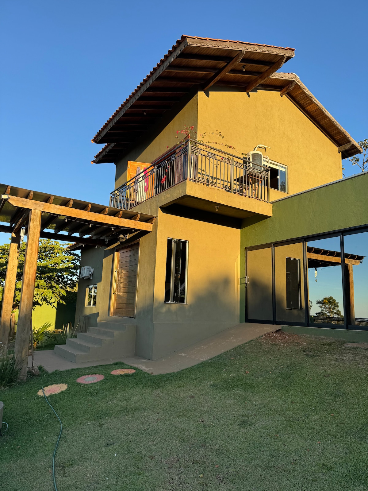 A two-story house is presented, featuring a wooden roof and a balcony adorned with greenery. Large glass windows provide abundant natural light, while a stone pathway leads to the entrance. The surrounding lawn is well-maintained, enhancing the outdoor space.