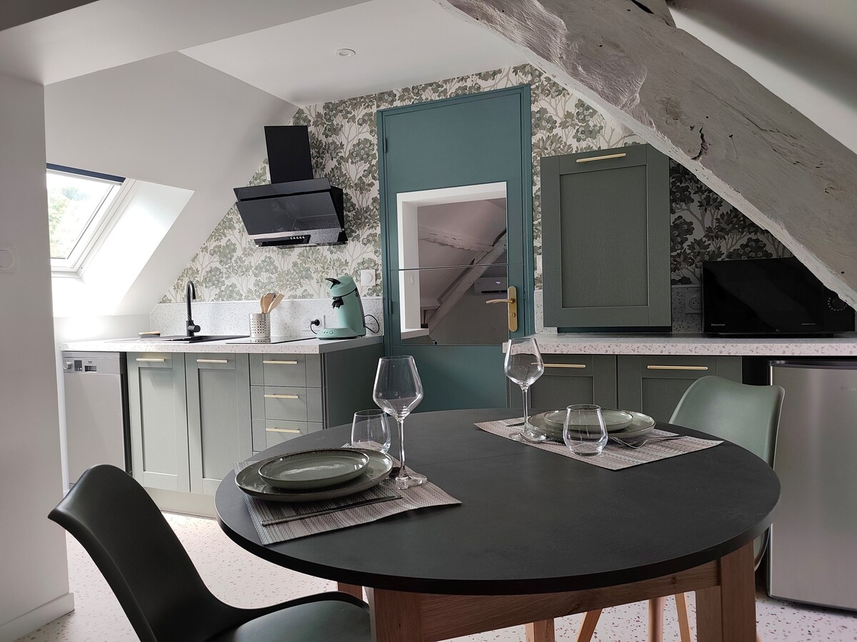 A modern kitchen area is visible, featuring a round dining table set for two with plates and glasses. Shelving units and a countertop are adorned with floral wallpaper. A stovetop is mounted under a sloped ceiling, complemented by appliances in soft green and stainless steel finishes.