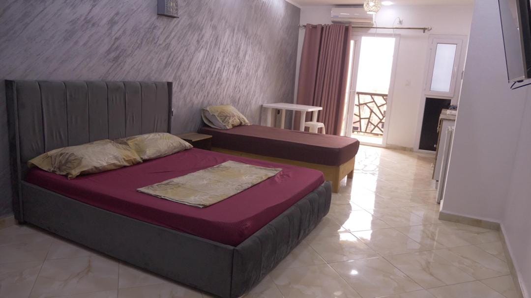 A spacious room features two beds, one with a comforter and decorative pillows. A small table is positioned nearby, providing additional seating. Natural light enters through a door leading to a balcony, while a television is mounted on the wall opposite the beds.