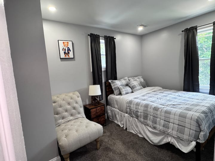 Útulná spálňa s manželskou posteľou, vzorovanou posteľnou bielizňou, tuftovaným kreslom, nočným stolíkom s lampou a veľkými oknami, ktoré prepúšťajú dostatok prirodzeného svetla.