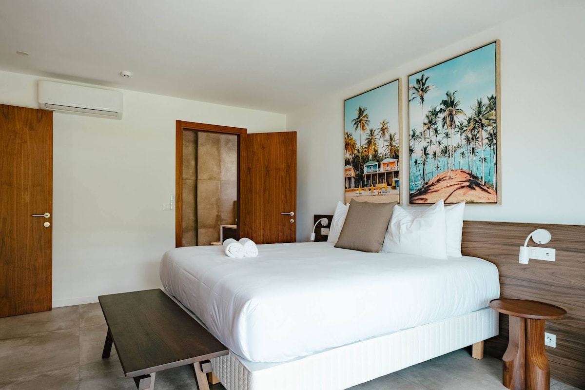 A spacious bedroom is depicted, featuring a comfortable bed with crisp white linens and a large wooden headboard. Two framed photographs of palm trees are hung on the wall, while natural light illuminates the room. A minimalist nightstand and wood accents enhance the modern decor.
