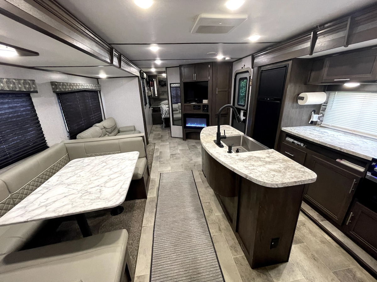 An inviting interior space features a well-equipped kitchen area with a curved marble countertop and dark cabinetry. A light-colored dining table with matching seating is positioned next to a cozy sofa. Large windows allow natural light while offering privacy with dark blinds.