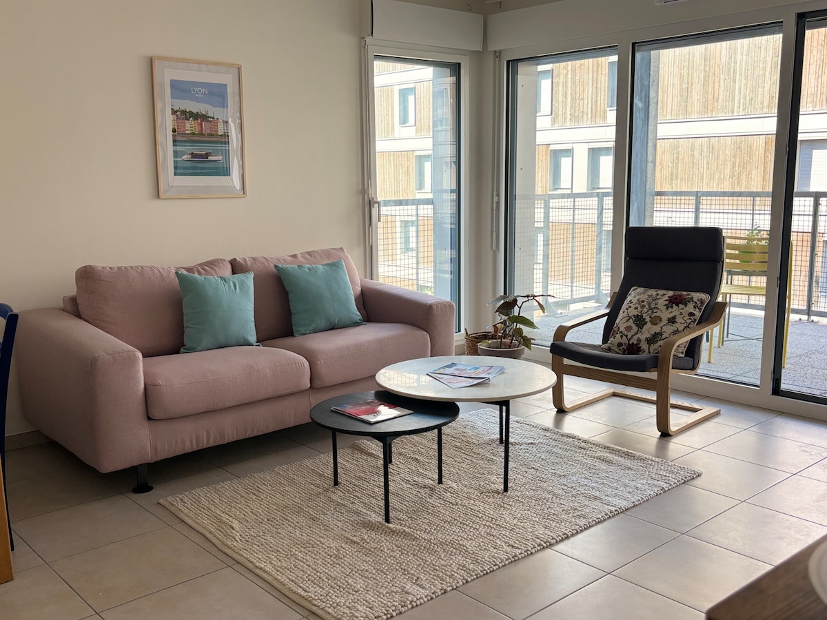 A modern living area is presented with a light pink sofa adorned with turquoise cushions. A wooden armchair features floral upholstery, accompanied by a woven area rug. Two round coffee tables are arranged on the tiled floor, and large windows allow natural light to fill the space.