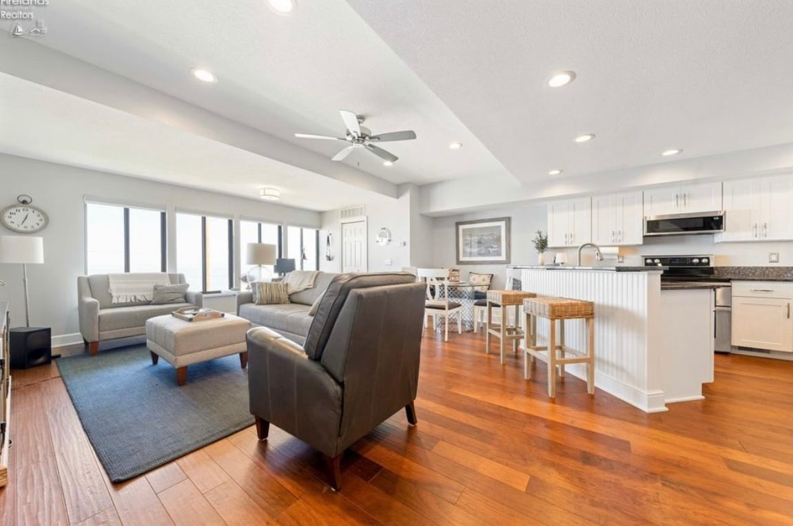 The open-concept living area features large windows offering lake views and natural light. Comfortable seating is arranged around a rug, with a dining area and modern kitchen visible in the background. A ceiling fan enhances airflow in the space, supported by warm wooden flooring.