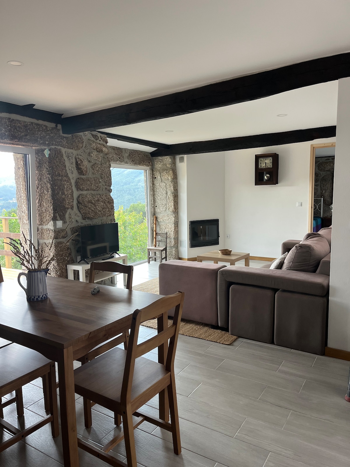 An open living space features stone walls and wooden beams. A cozy sofa is positioned near large windows, offering views of the lush landscape. A dining table with four chairs is placed nearby, and a fireplace adds an element of comfort.