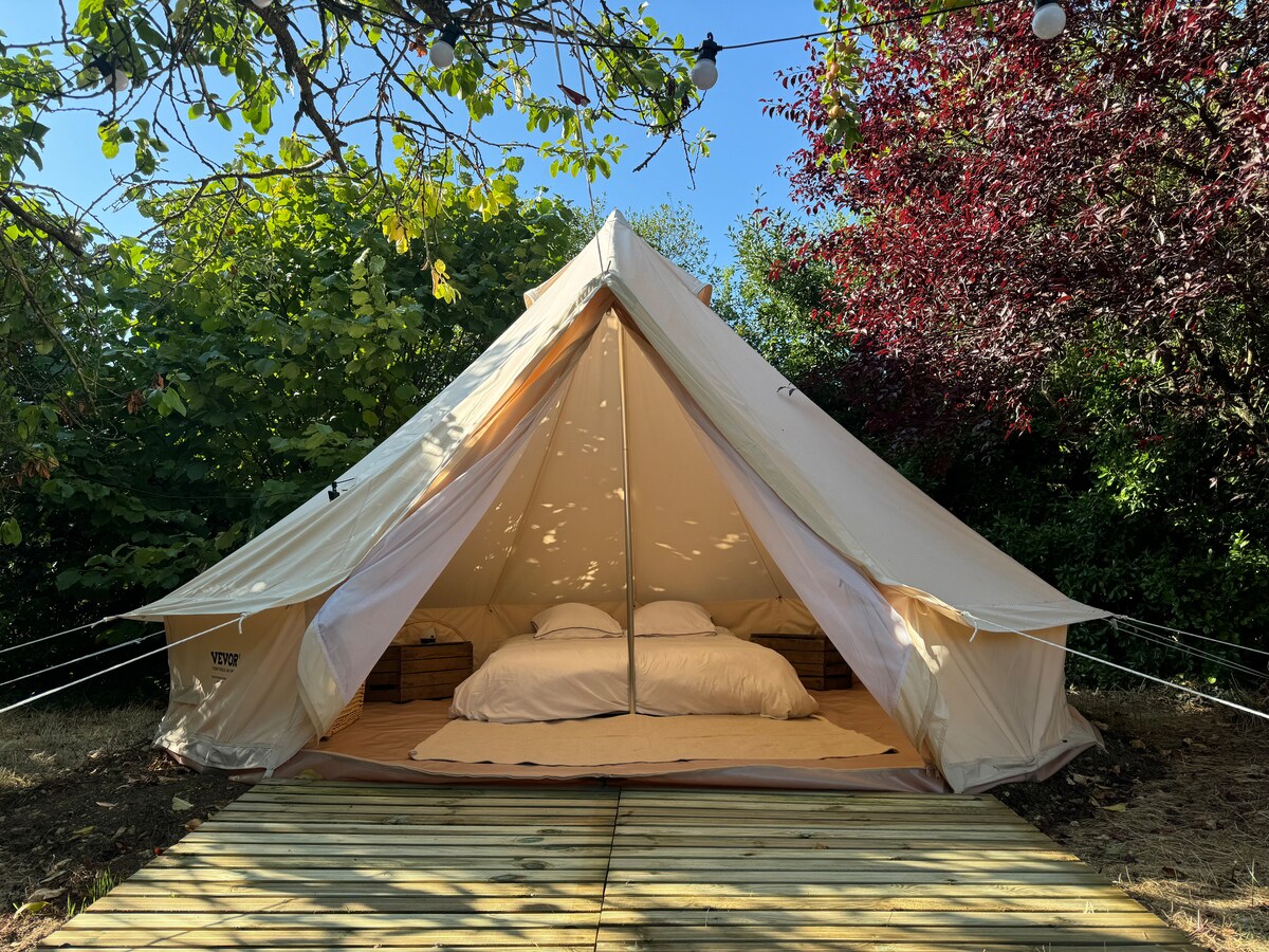 A spacious tipi is situated on a wooden deck surrounded by greenery. The entrance is open, revealing a comfortable double bed with white bedding. Natural light filters through the canopy of trees, enhancing the serene outdoor setting.