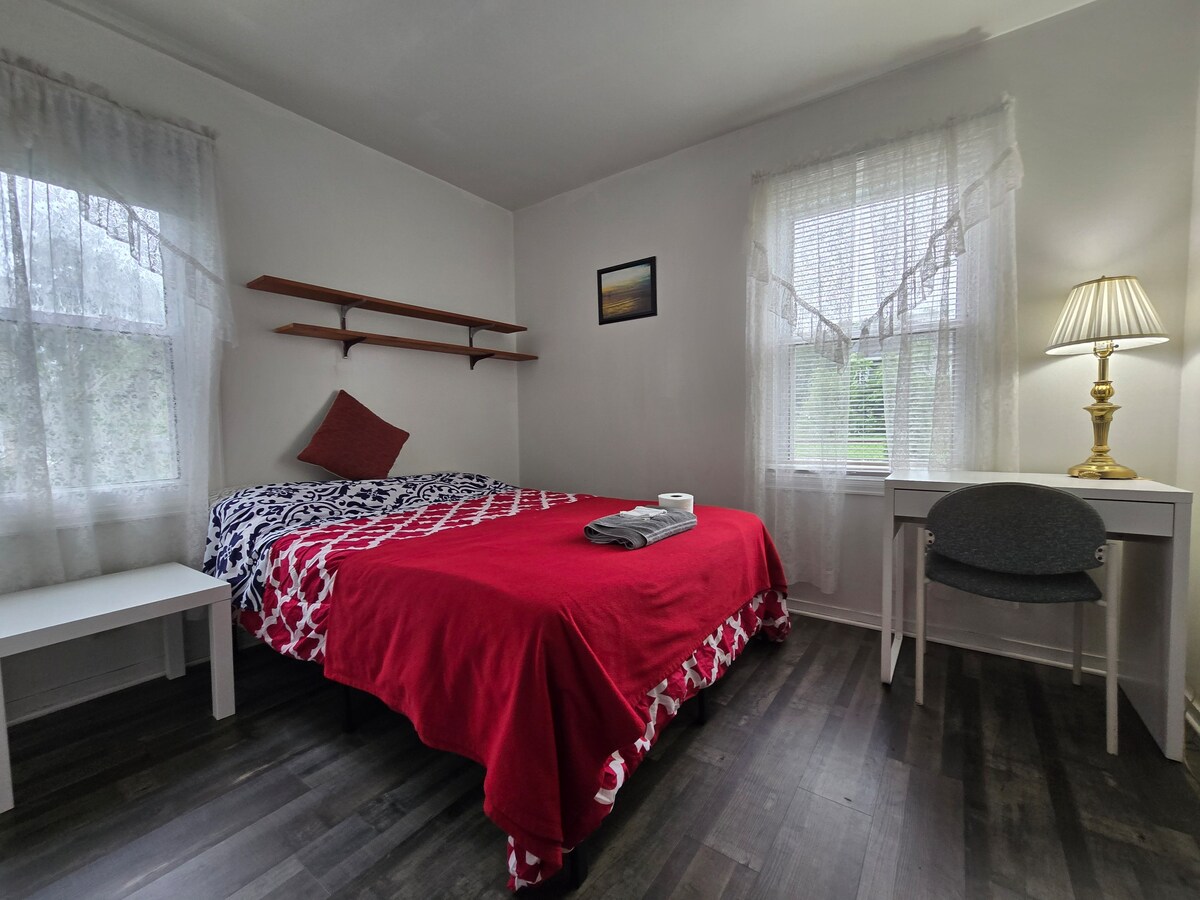 A room features a spacious bed covered in a red and white patterned quilt. Natural light enters through two windows with sheer curtains. A small desk with a lamp and a simple chair is positioned next to the bed, along with a shelf mounted on the wall.
