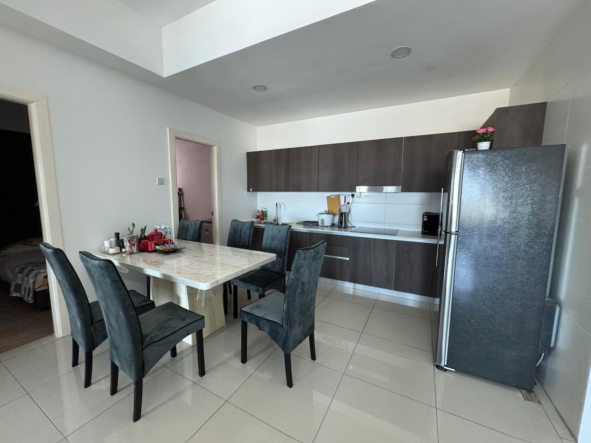 A fully equipped kitchen and dining area is displayed, featuring a large dining table surrounded by several dark upholstered chairs. Modern cabinetry is shown alongside stainless steel appliances, including a refrigerator. Natural light brightens the space through a window, creating a welcoming environment.