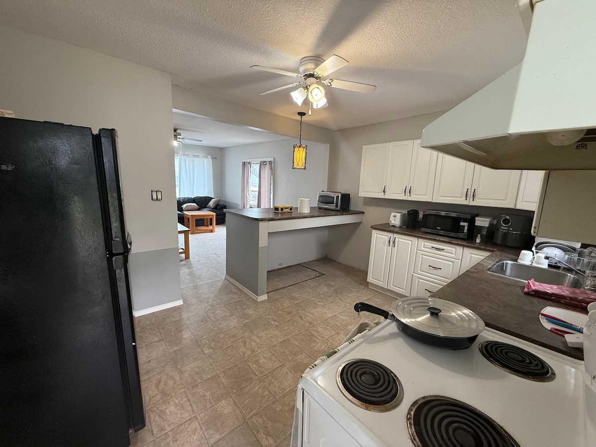 A kitchen area features a stove and an adjacent countertop with various appliances, including a microwave and coffee maker. The open layout allows for a view into the living space beyond, where light enters through large windows, creating a bright atmosphere.