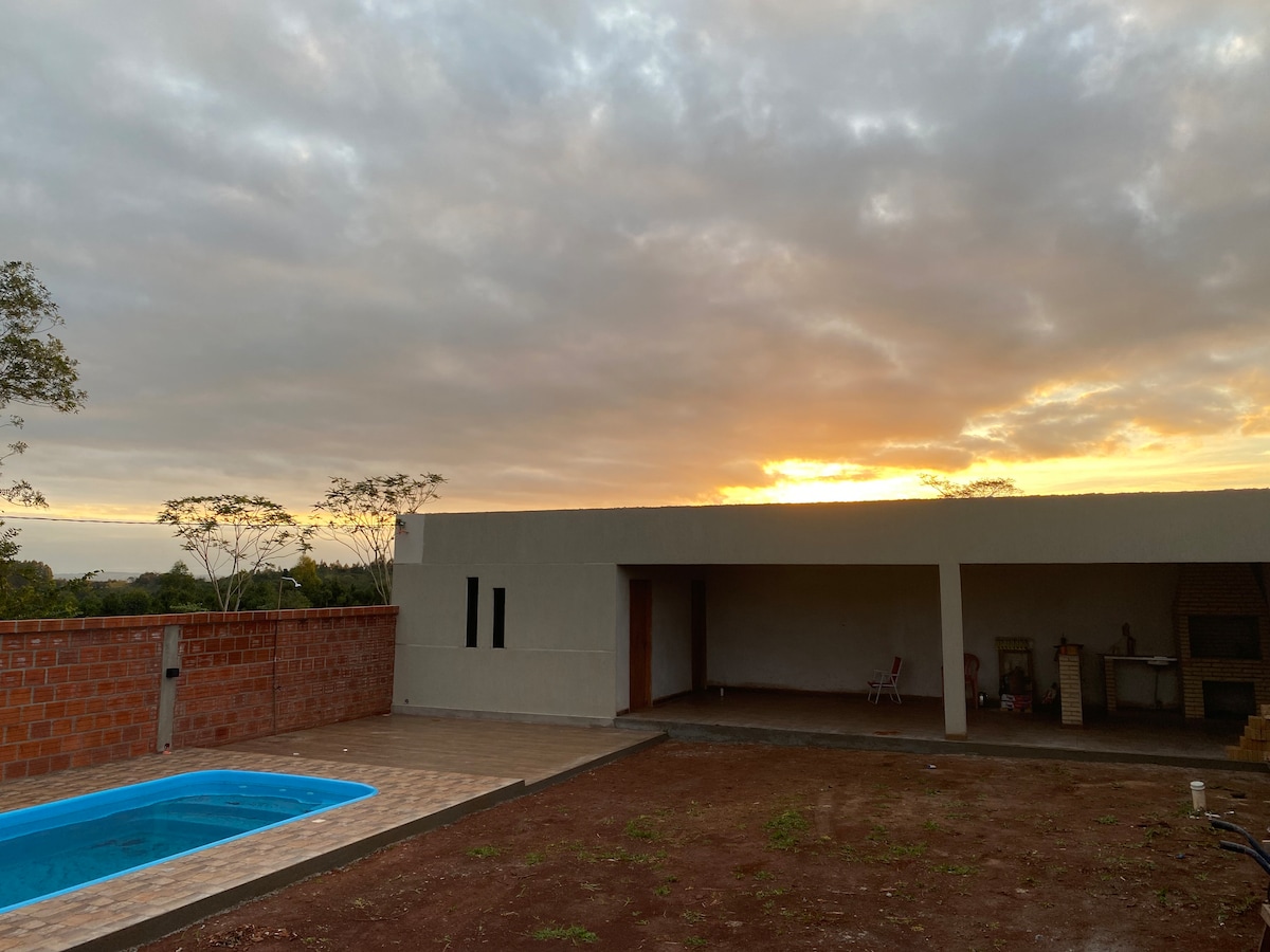 A tranquil outdoor space is depicted, featuring a small swimming pool with a patterned deck. A single-story building is visible nearby, with a covered area that offers kitchen amenities. The sky above displays soft clouds, and the sunset casts a warm glow over the setting.