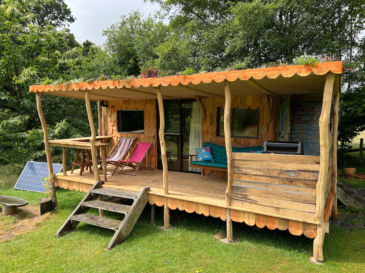 A spacious wooden veranda extends from the Shepherd's Hut, featuring colorful seating options, and surrounded by lush greenery. Solar panels are visible, showcasing the eco-friendly design. Steps lead to the entrance, inviting guests onto the outdoor space for relaxation or meals.