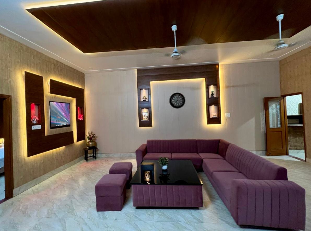 A spacious living area features a large, plush sectional sofa arranged around a central glass coffee table. Soft lighting accentuates the designer decor, with wall-mounted shelves displaying decorative items. A television is positioned on the wall, enhancing the functionality of the space.