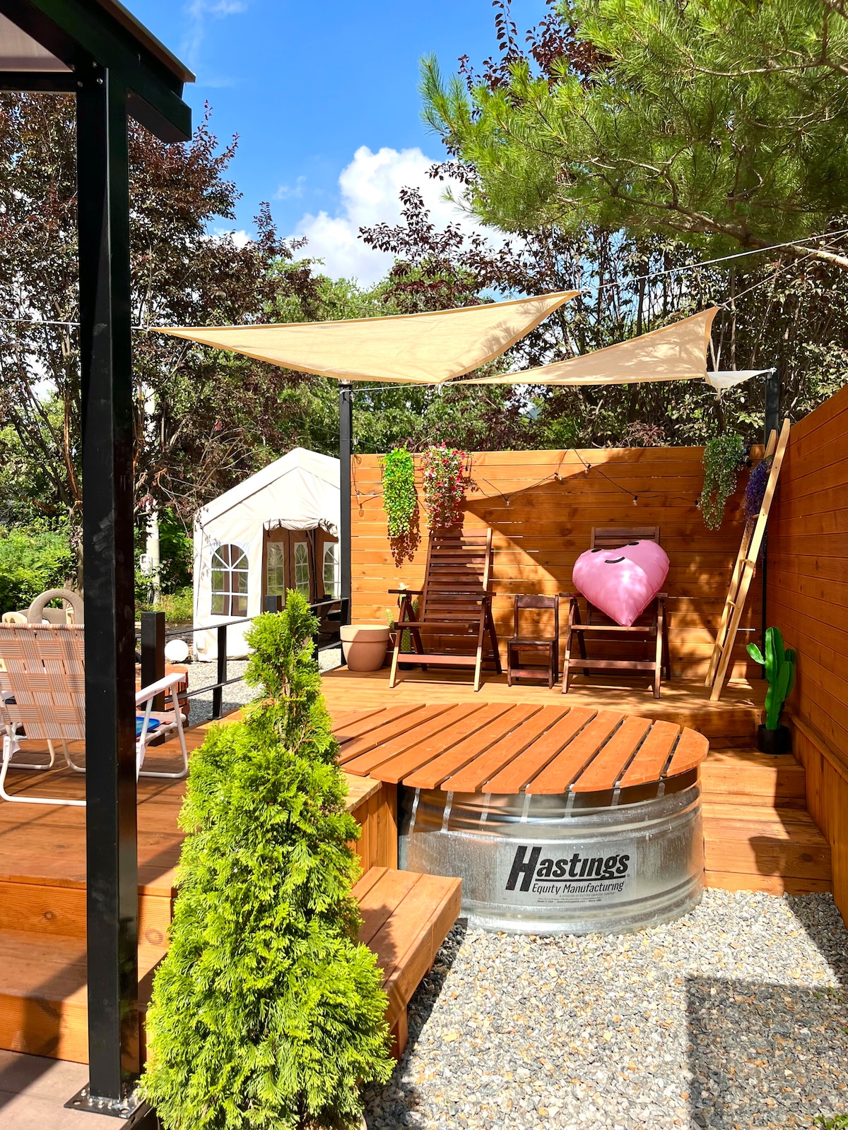 A tranquil outdoor area features a circular metal hot tub surrounded by a wooden deck. Two reclining chairs are positioned nearby, complemented by potted plants and a decorative heart. Shade sails provide coverage, enhancing the cozy environment amid lush greenery.
