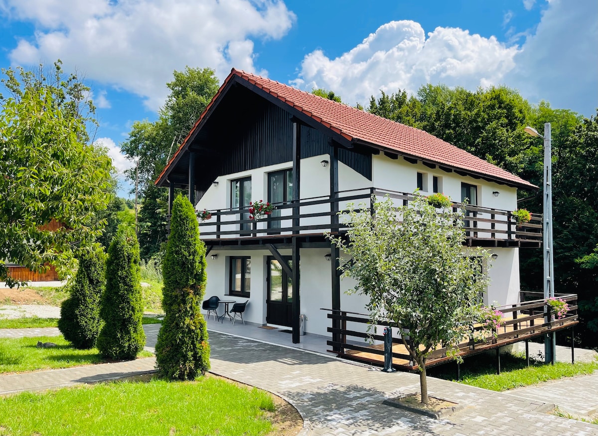 A spacious cabin is visible against a backdrop of greenery, featuring a two-story structure with a red-tiled roof. A wooden balcony with planters adorns the front, complemented by neatly paved pathways and landscaped areas with shrubs and trees.