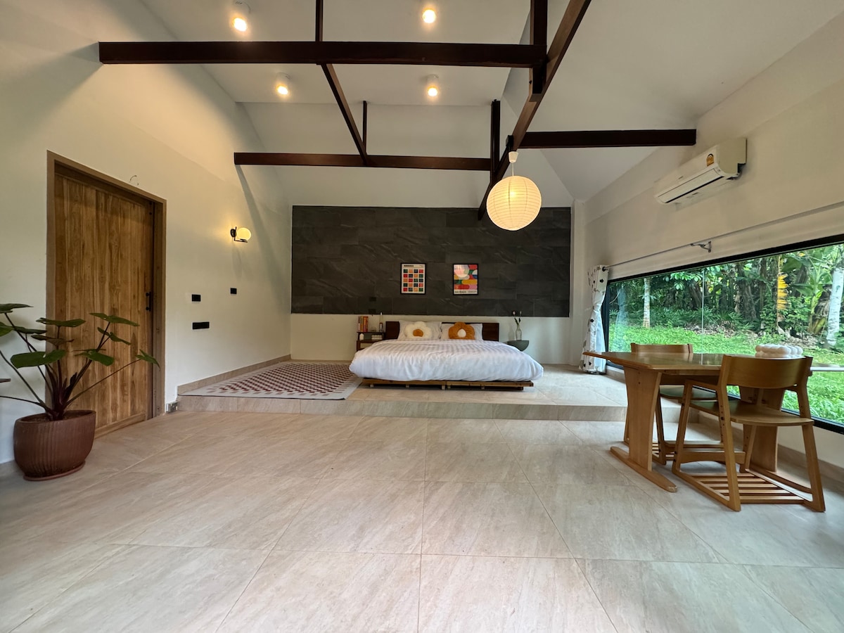 An open and airy cabin interior is displayed, featuring a spacious bed positioned against a slate accent wall. A dining area with two chairs and a small table is visible, alongside a large window that offers views of the surrounding greenery.