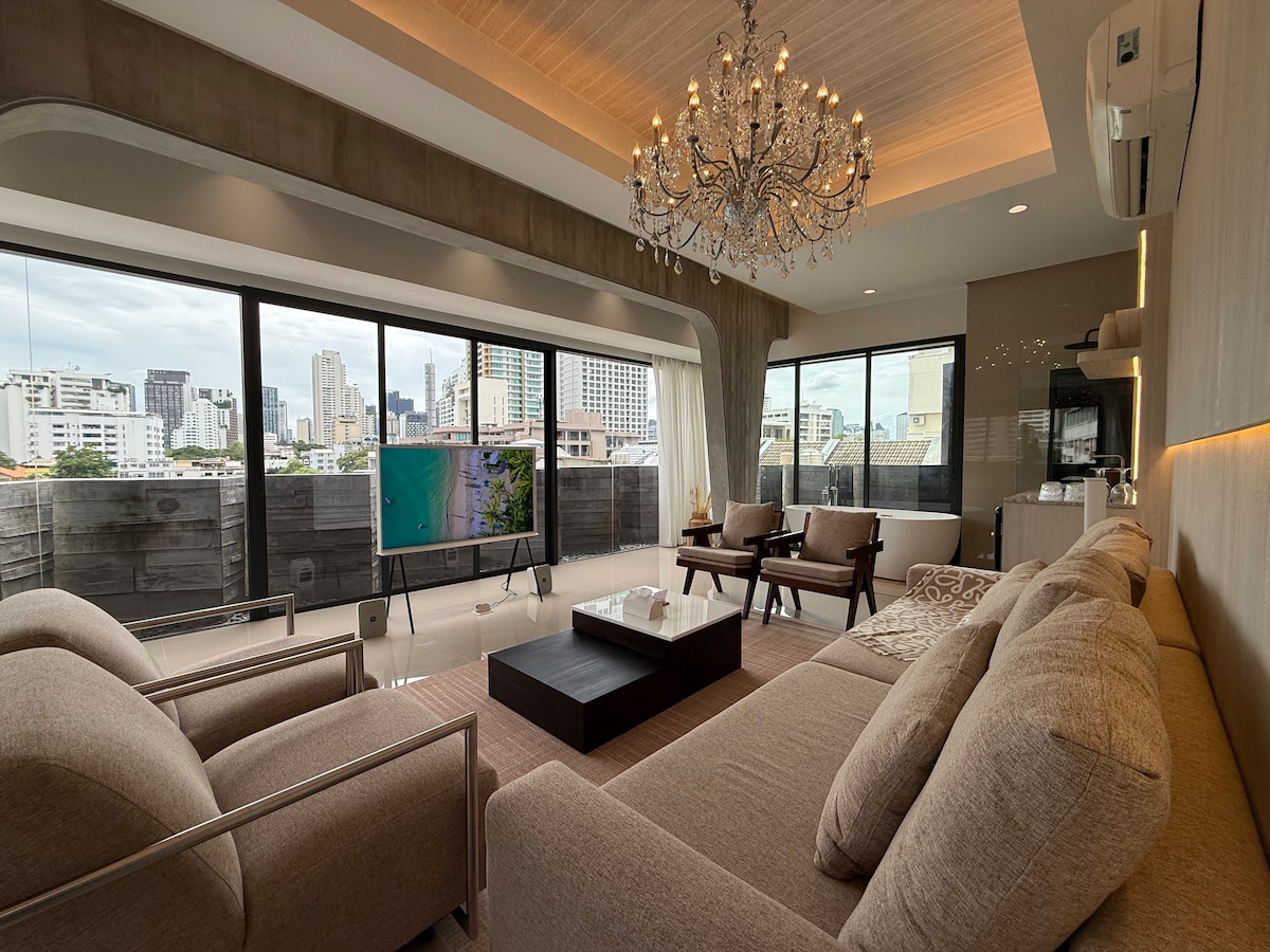 A spacious living room features large floor-to-ceiling windows that offer panoramic views of Bangkok City. Elegant furnishings include a cozy sectional sofa and two armchairs, complemented by a central coffee table. A stylish chandelier hangs from the ceiling, enhancing the modern atmosphere.