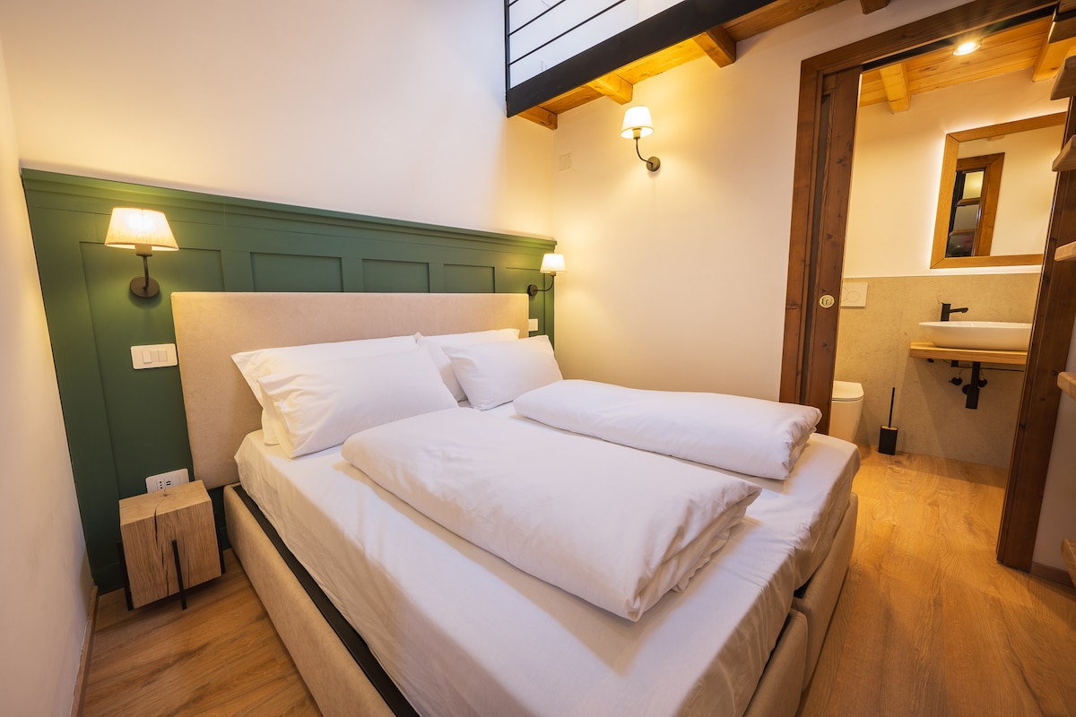 A well-lit attic room features a spacious bed with crisp white bedding. Soft bedside lamps provide warm lighting, while wooden beams enhance the room's character. A small wooden nightstand sits beside the bed, and a bathroom can be seen through an open door.