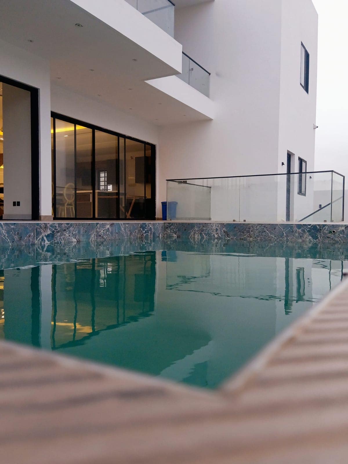 A tranquil infinity pool reflects the modern architecture of the adjacent building. The water's surface appears still, with a smooth edge blending into the surrounding deck. Large glass doors provide glimpses of the open interior space, enhancing the sense of connection between indoor and outdoor areas.