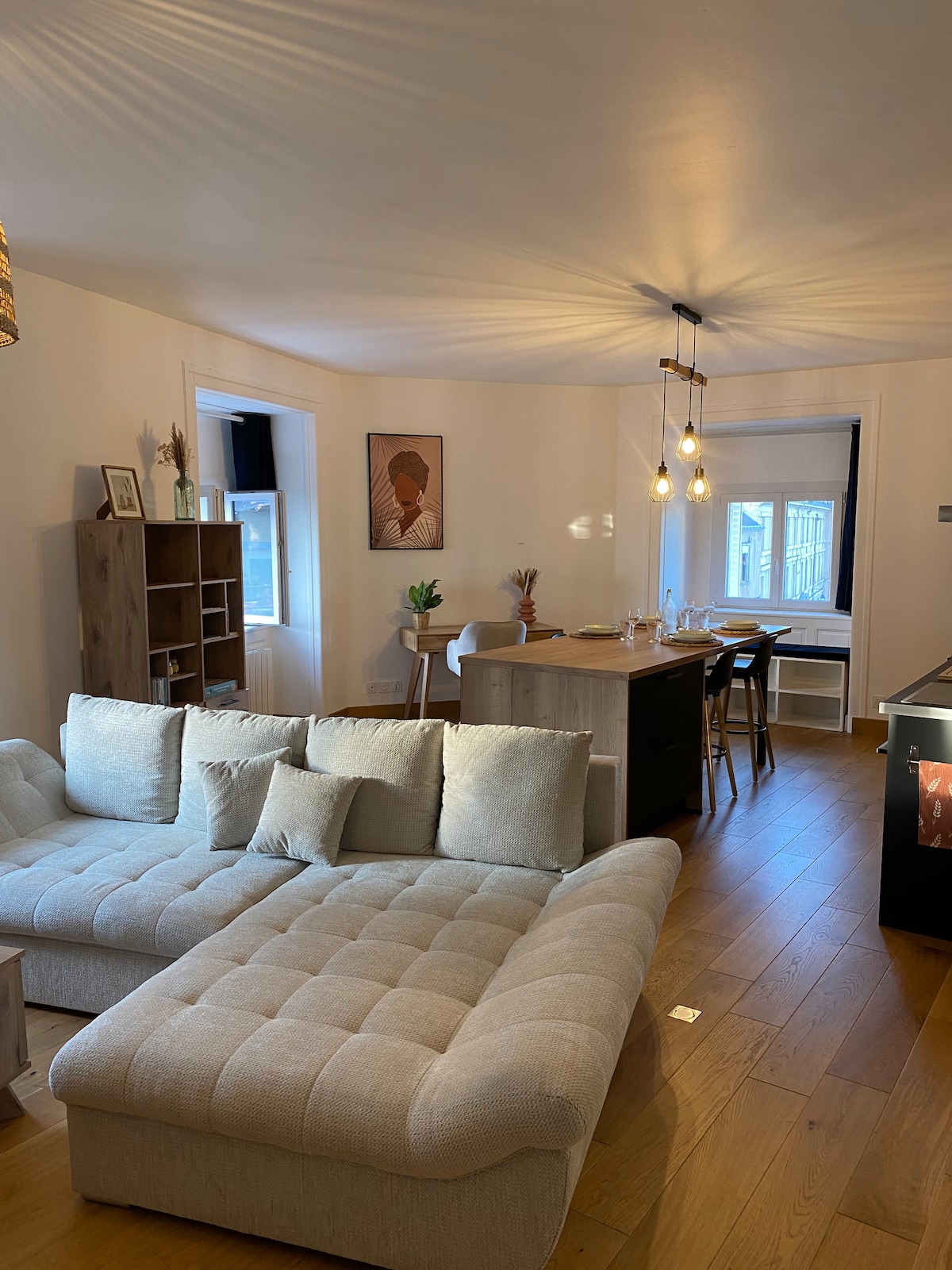 The open-plan living area showcases a light-colored sectional sofa with multiple cushions, inviting relaxation. A dining table with chairs is visible in the background, illuminated by pendant lighting. A window provides natural light, and tasteful artwork adds character to the space.