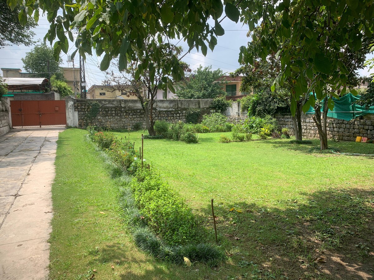 A spacious private lawn is surrounded by lush greenery, featuring well-maintained grass and a variety of plants. The area is framed by a stone wall and trees, providing a serene environment for relaxation. A paved pathway leads from the lawn to the entrance gate.