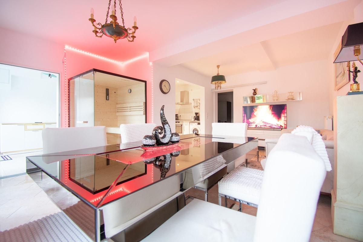 A modern dining area features a sleek mirrored table surrounded by white chairs. Soft pink lighting highlights the space, where a decorative centerpiece rests on the table. An electric fireplace can be seen in the background, providing a cozy ambiance.