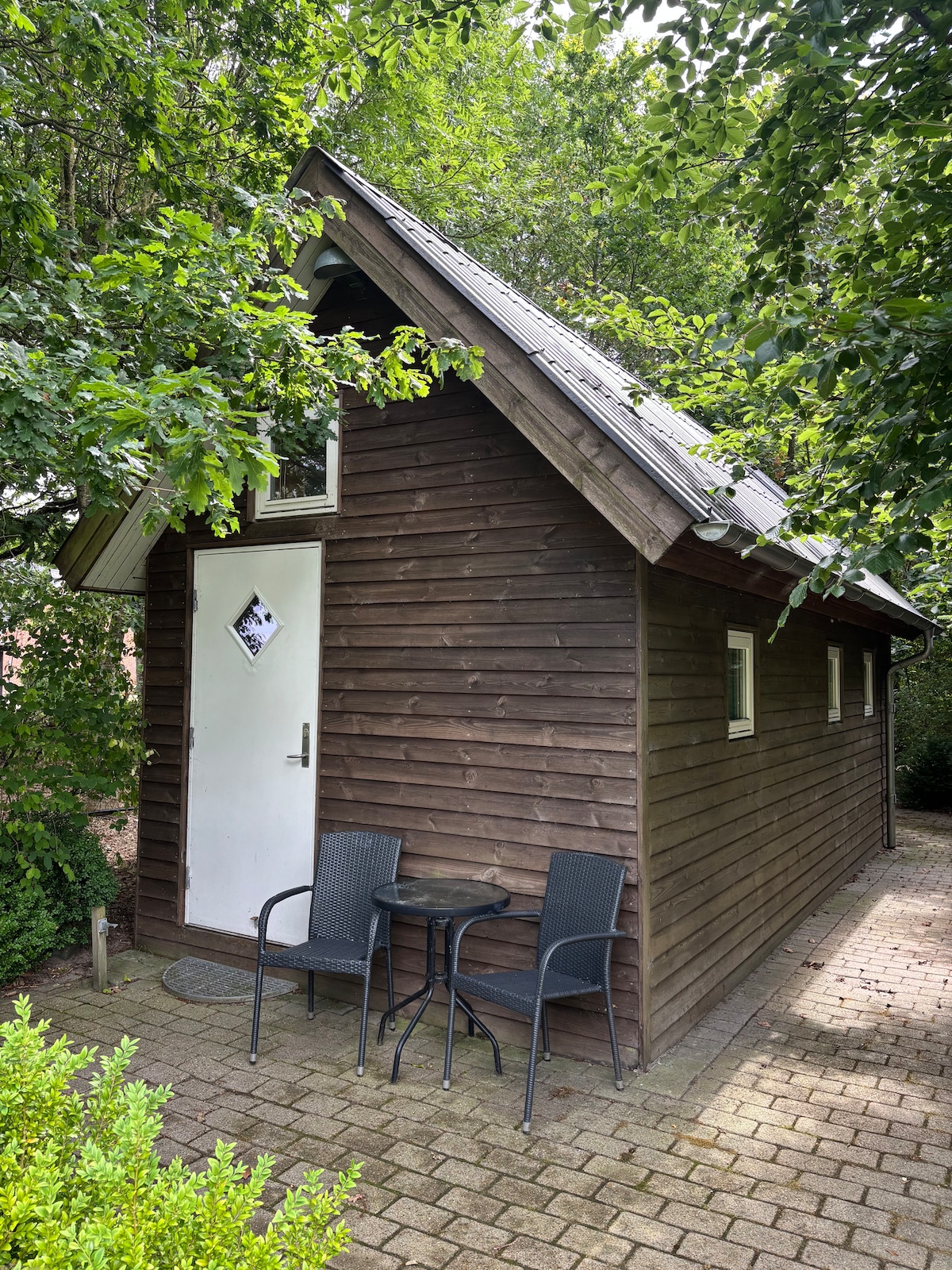 A wooden cabin features an angled roof and a welcoming entrance door. Two black chairs and a small table are positioned outside, surrounded by lush greenery. The path is paved with rectangular stones, leading to the door, which is marked with a unique sign.