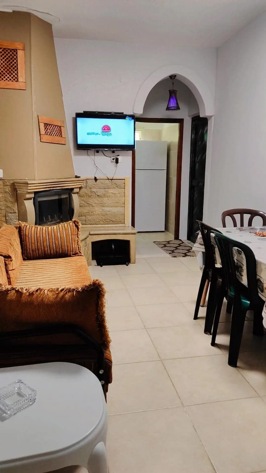 A cozy living area is highlighted by a beige sofa adorned with striped cushions. A television is mounted on the wall, next to a doorway leading to a kitchen with a refrigerator. A dining table with several chairs occupies the space, providing a welcoming atmosphere.