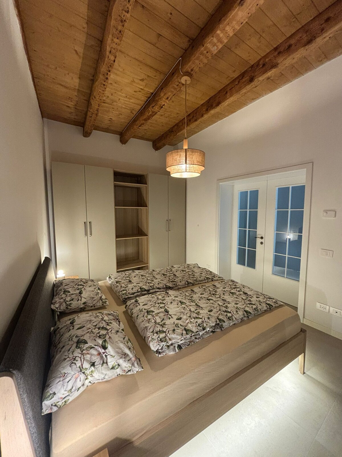 A bright bedroom features a spacious bed adorned with floral-patterned bedding. Light wood accents are present in the ceiling beams and furniture. A wardrobe is partially open, showing storage space, and double glass doors lead to another area, allowing natural light to filter in.
