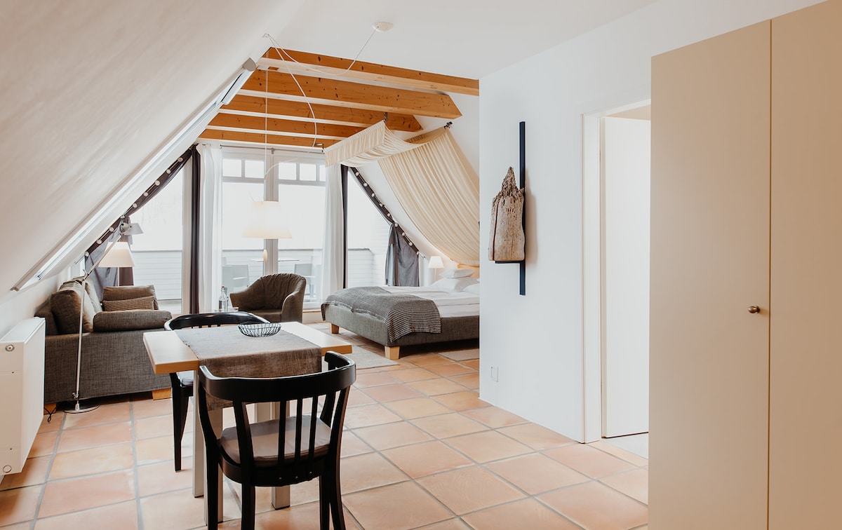 The studio's interior features a spacious layout with a large double bed and a separate single bed. A dining table with black chairs is positioned nearby, and a cozy seating area includes a sofa. Natural light fills the room through large windows, showcasing warm wooden beams.