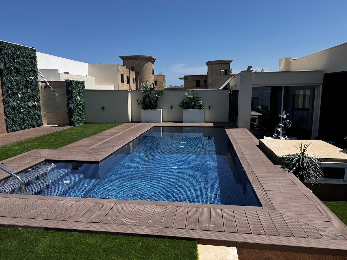 An outdoor pool is situated on a wooden deck surrounded by lush greenery and minimalistic white planters. The water reflects the clear blue sky, and modern architecture is visible in the background, creating a serene and private outdoor space.