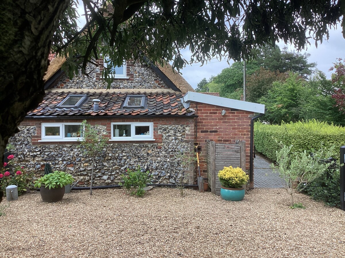 The exterior of a charming thatched cottage is displayed, featuring a mix of flint and brick walls. A pebble-strewn area surrounds the cottage, with potted plants and greenery contributing to a natural landscape. Windows are visible, reflecting the surrounding foliage.