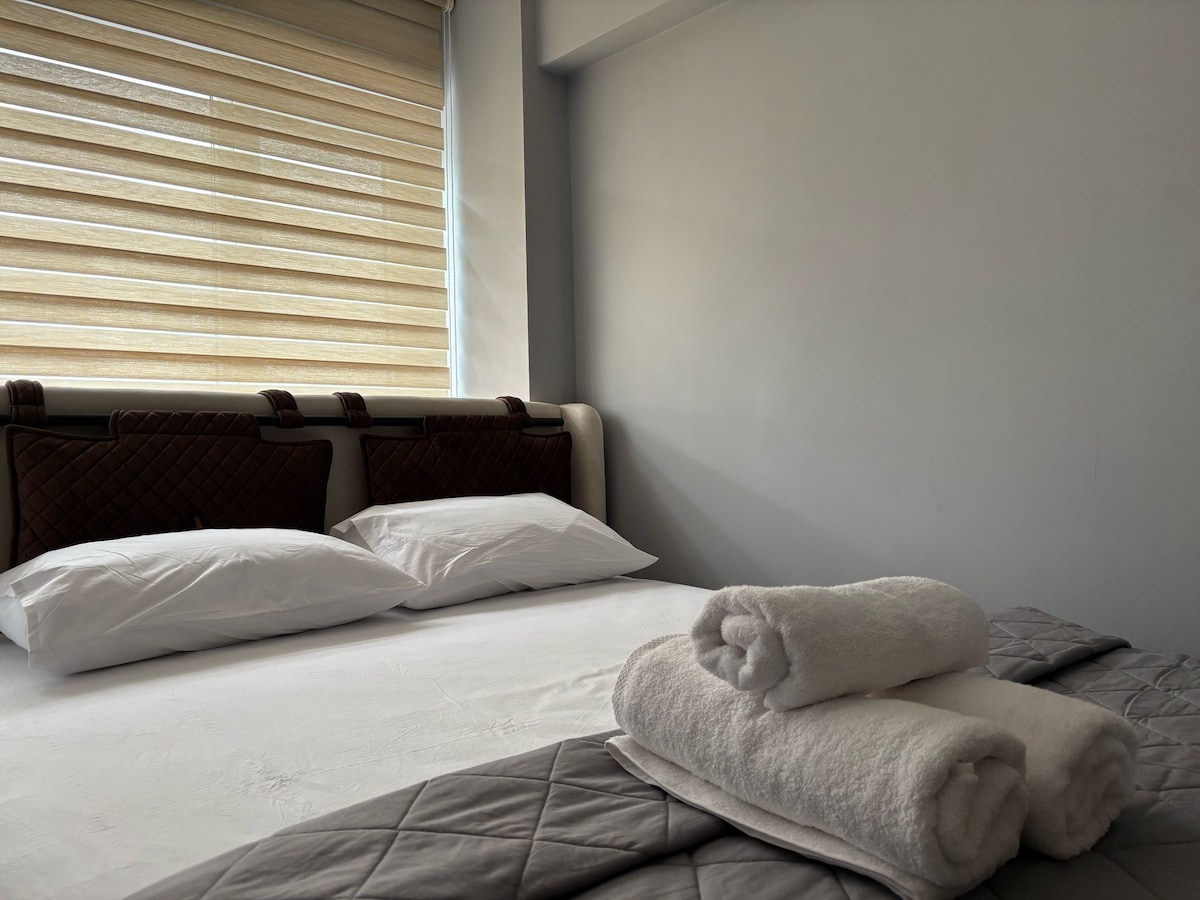A spacious bed is visible, consisting of two white pillows and a neutral-colored blanket. Rolled towels are neatly arranged on the bed's edge. Soft, natural light filters in through a partially open window with textured shades, contributing to a calm ambiance.