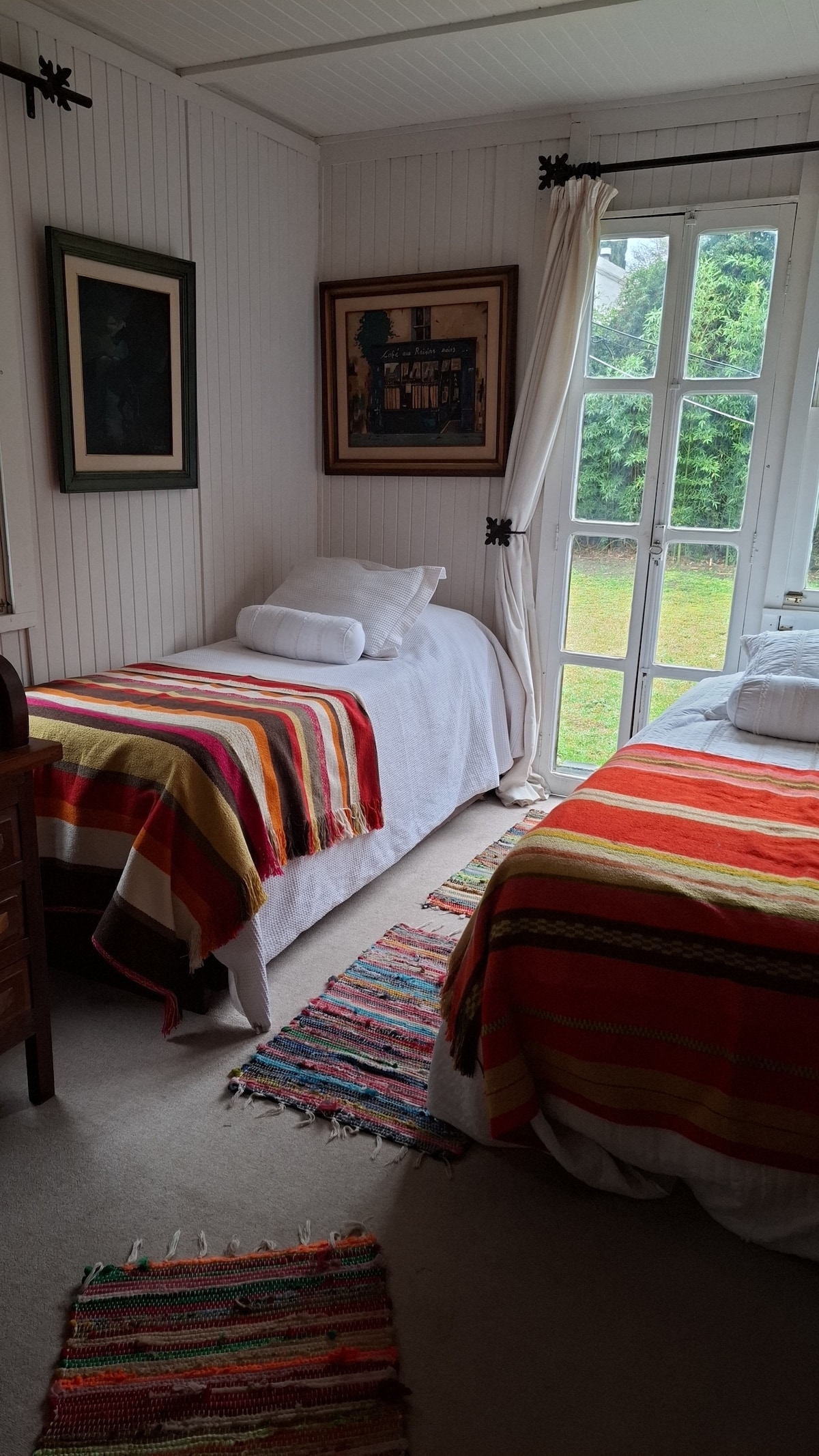 A bright bedroom features two twin beds dressed in white with colorful striped blankets. Natural light enters through large windows, revealing a view of the greenery outside. A decorative, patterned rug lies on the carpeted floor, adding a touch of warmth to the space.