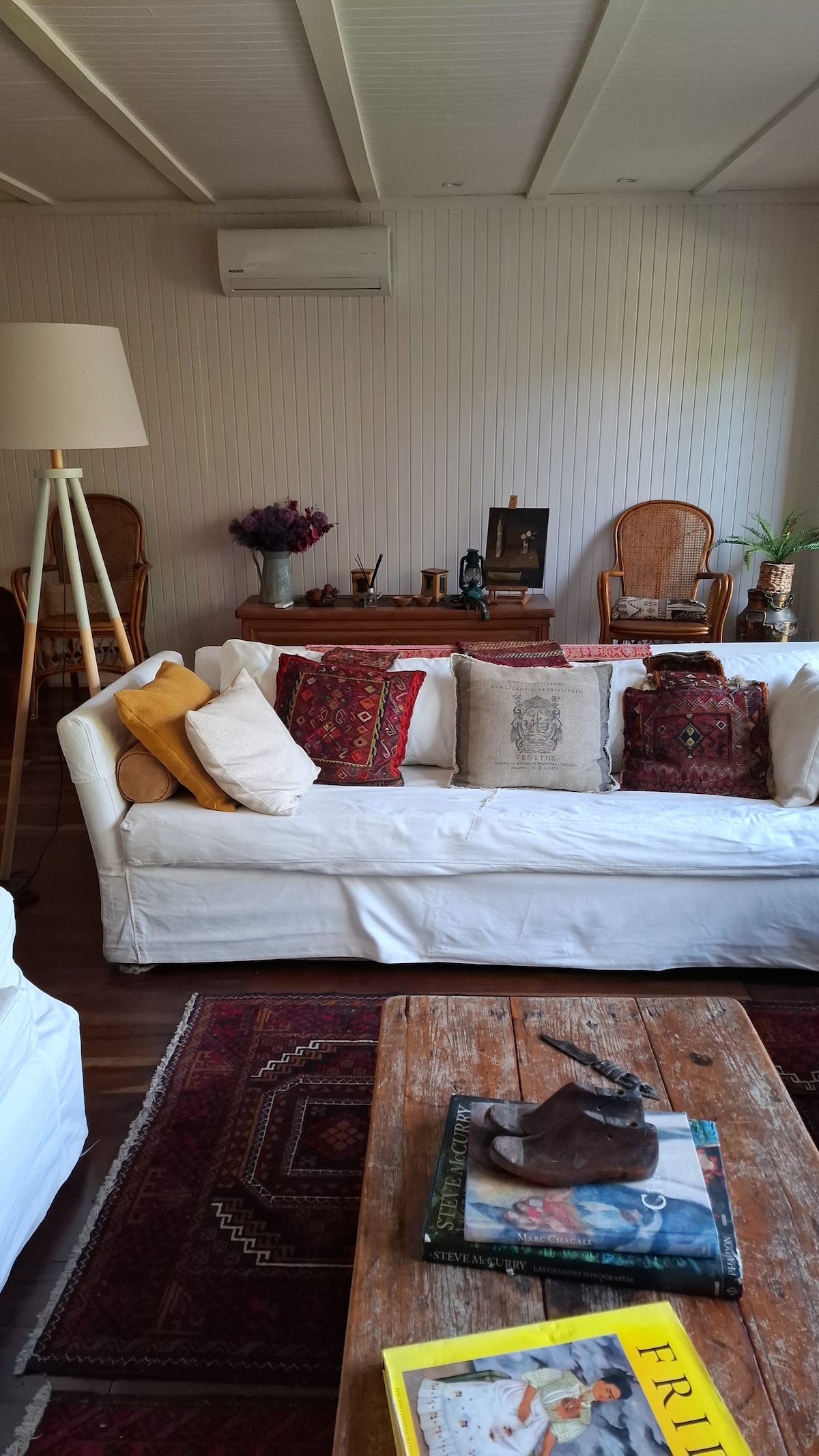 A cozy living area features a white sofa adorned with decorative pillows, set against a backdrop of neutral walls. A rustic wooden coffee table is placed centrally, surrounded by an assortment of books. A lamp and decorative seating add warmth to the space.