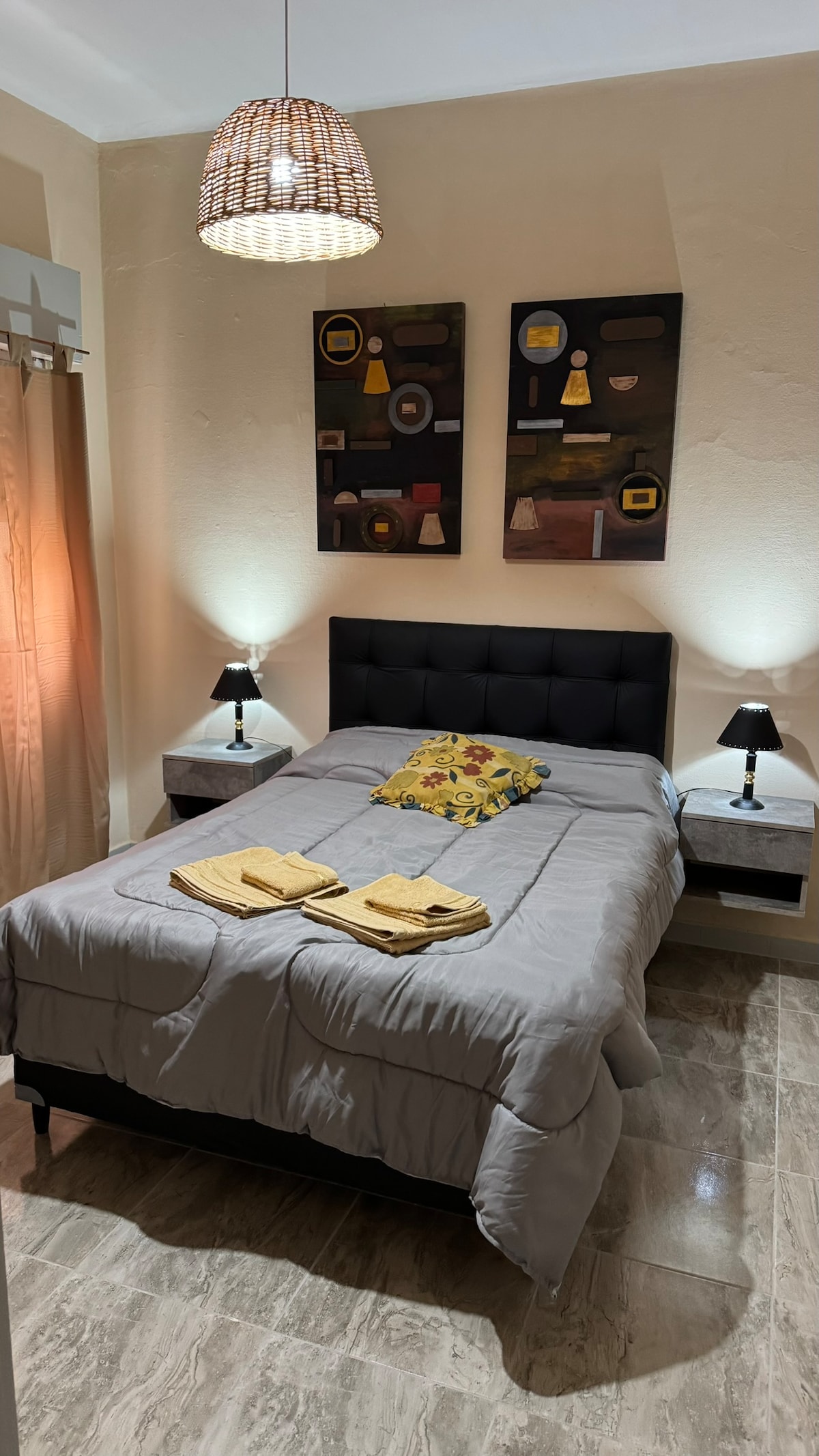 A sleeping area is presented with a spacious bed, adorned with a gray coverlet and a decorative pillow featuring floral patterns. Two bedside tables, each with lamps, are positioned on either side. Wall art adds a touch of character, while warm lighting creates a cozy ambiance.