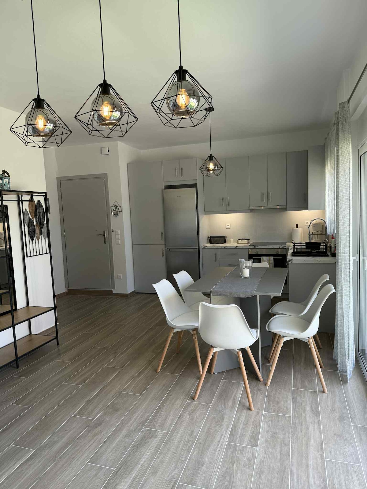A modern kitchen and dining area is presented, featuring a sleek table with four white chairs arranged around it. Pendant lights hang above the table, illuminating the space. A stainless steel refrigerator and a fully-equipped kitchen are visible in the background, enhancing functionality.
