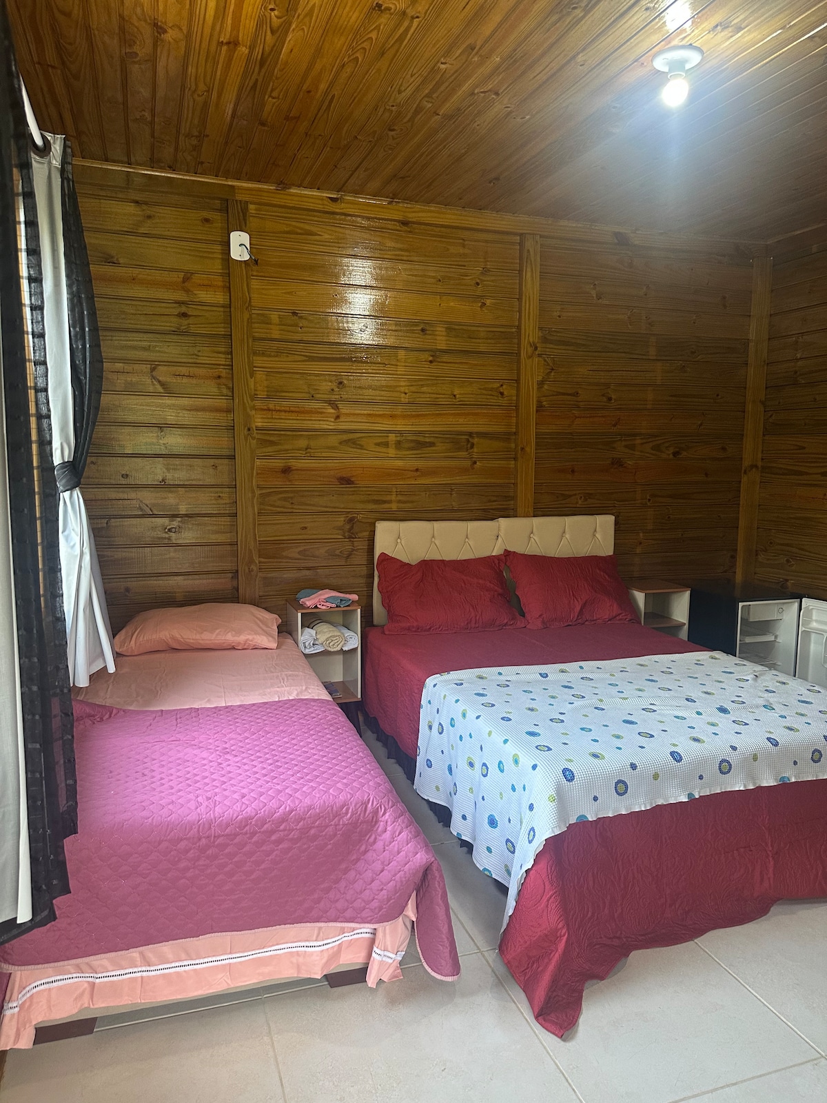 A sleeping arrangement features a spacious bed and an additional single bed, both adorned with soft bedding. The wooden walls create a warm ambiance, while natural light is filtered through dark curtains, enhancing the cozy atmosphere of the room.