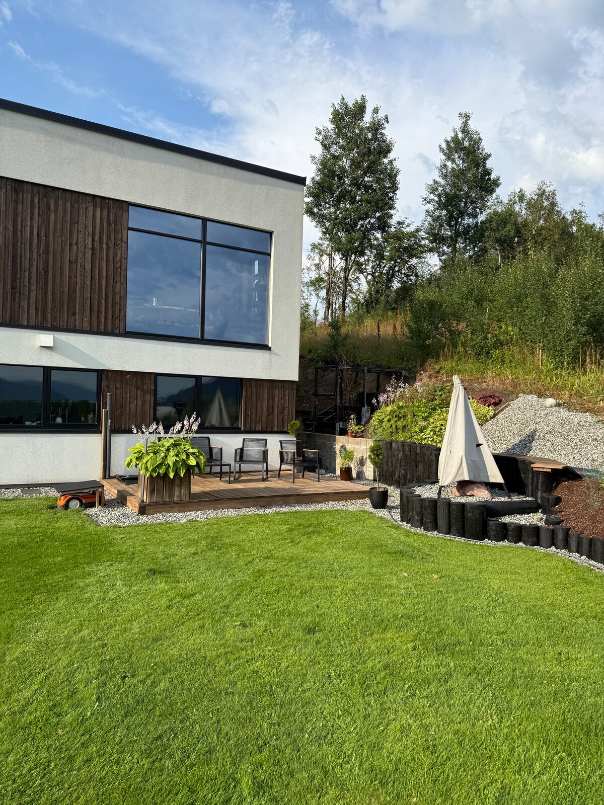The exterior of a modern house is presented, featuring large windows that invite natural light. A wooden deck is equipped with chairs, while a grassy area includes landscaped elements. Surrounding greenery and a small decorative feature enhance the outdoor ambiance.