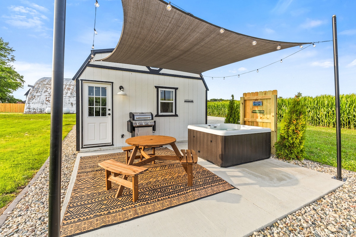 An outdoor patio area is showcased with a round picnic table and two benches placed on a patterned rug. A hot tub is situated nearby, with a grill positioned against the cabin wall. Decorative string lights are draped above, adding a relaxed ambiance.