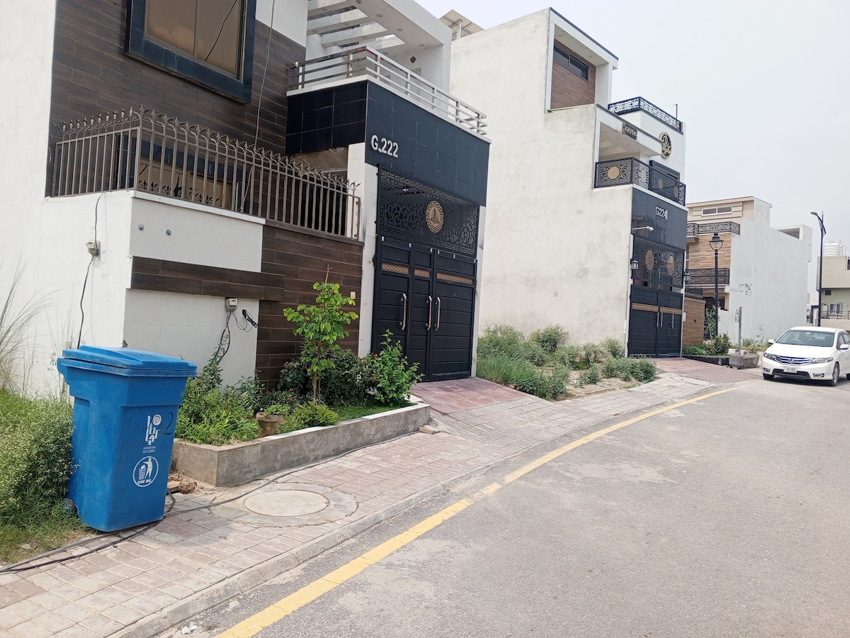 A view of two contemporary buildings located along a paved street, featuring dark entrances with circular motifs. A blue waste bin is positioned next to a small green plant. The surroundings include low shrubs and a smooth road curving into the distance.