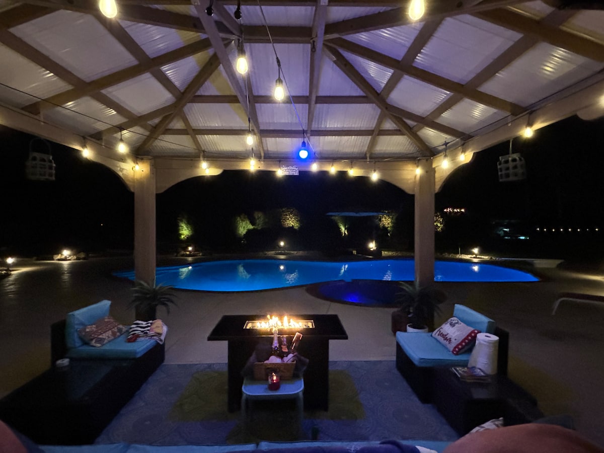An outdoor gazebo is illuminated by string lights, creating a cozy atmosphere. A fire pit is surrounded by comfortable seating, with a view of a pool reflecting soft blue lights in the background. Lush landscaping is visible, enhancing the serene setting at night.