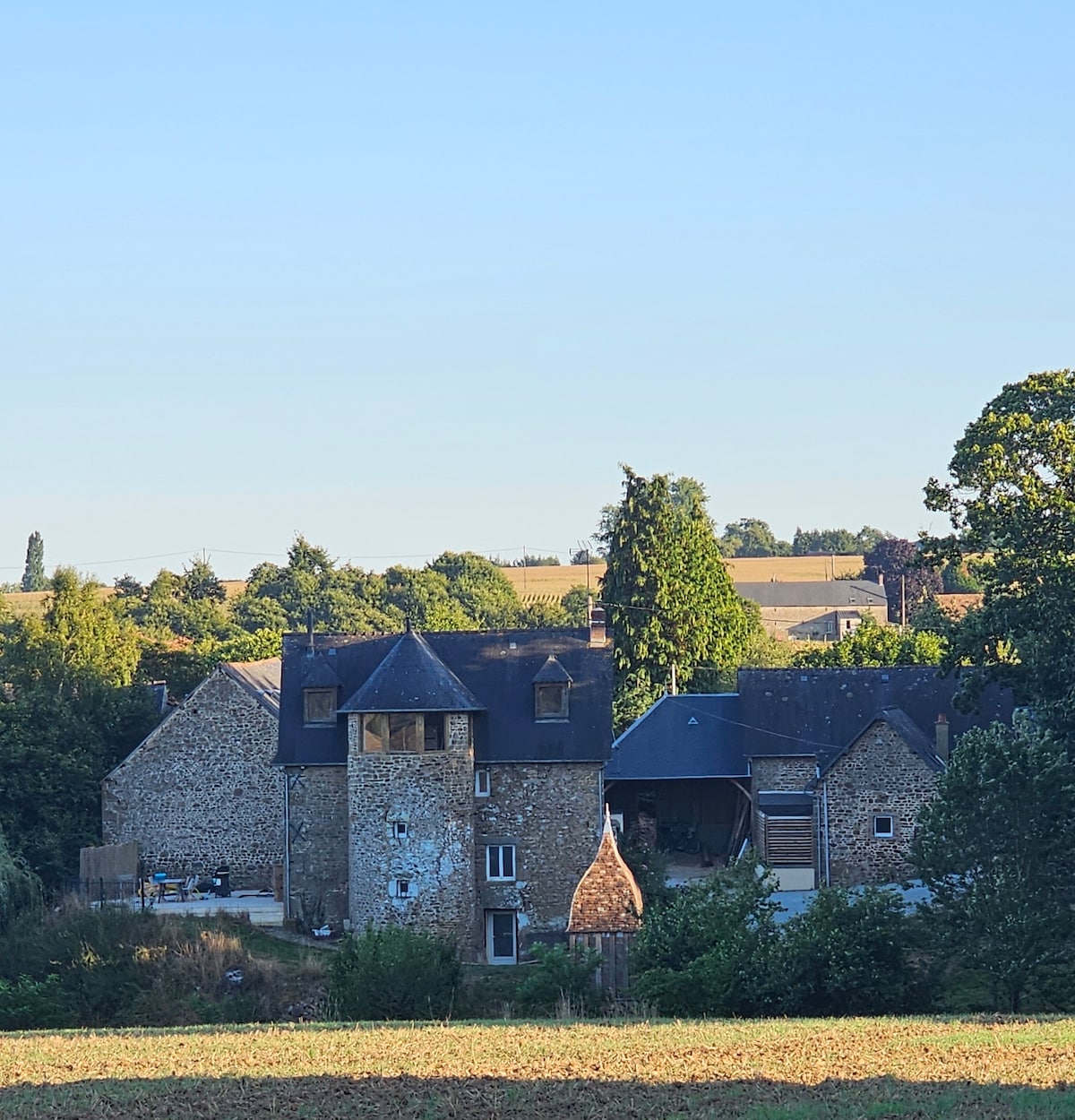 A charming stone farmhouse is observed from a distance, characterized by its varied rooflines and rustic architecture. Surrounding greenery and distant fields complement the structure, which reflects a blend of historical charm and modern amenities.