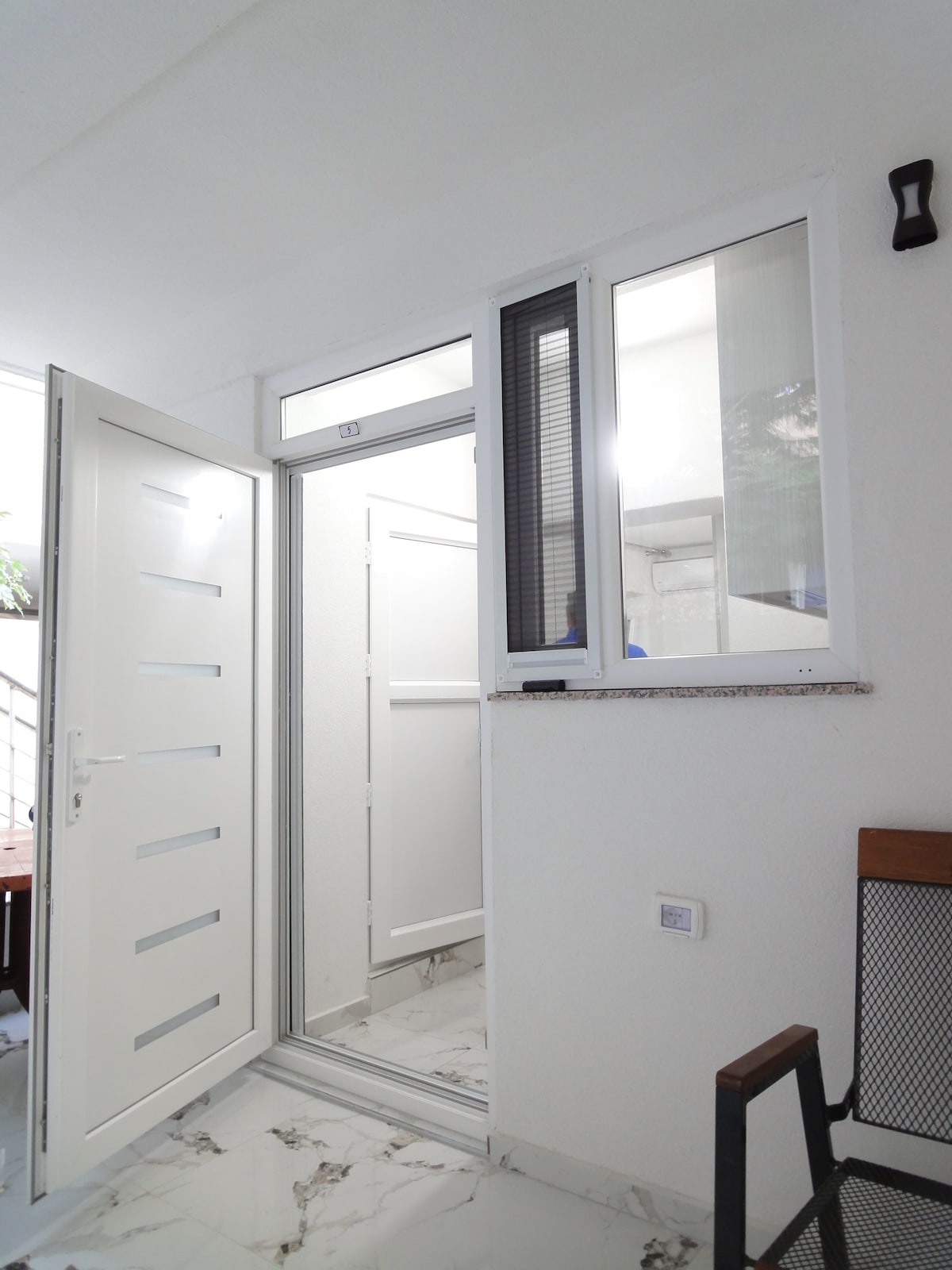An entrance area features a modern white door with horizontal panels. Adjacent, a large window allows natural light to brighten the space. Smooth marble flooring complements the clean design, contributing to a fresh and open atmosphere.