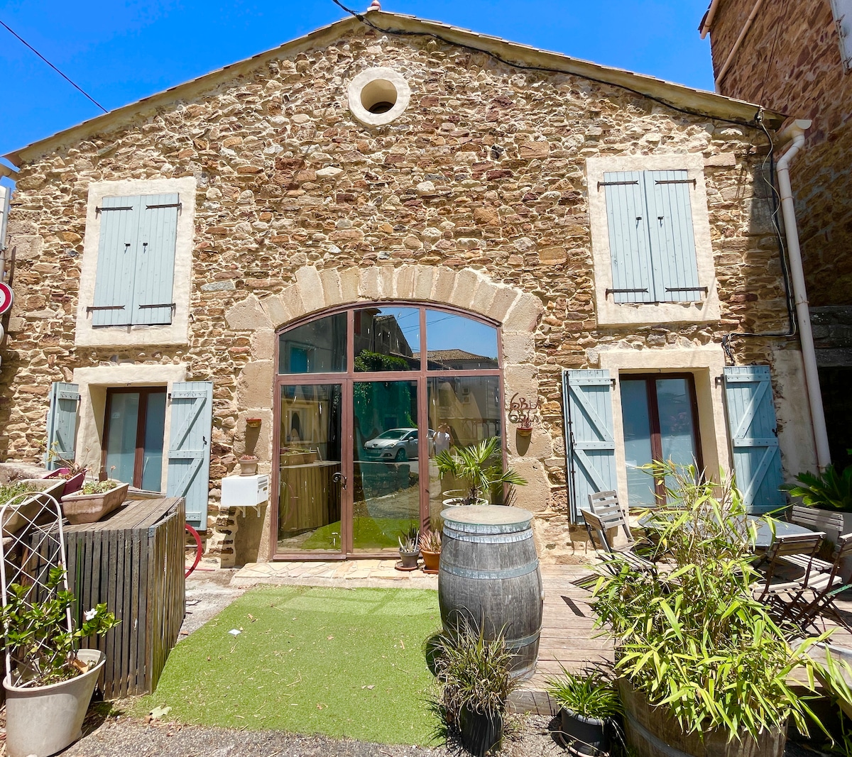 The stone exterior of the house is complemented by light blue shutters and an arched window, allowing natural light to fill the interior. Potted plants and outdoor seating are positioned on a stone pathway, adding a touch of greenery to the inviting entrance.