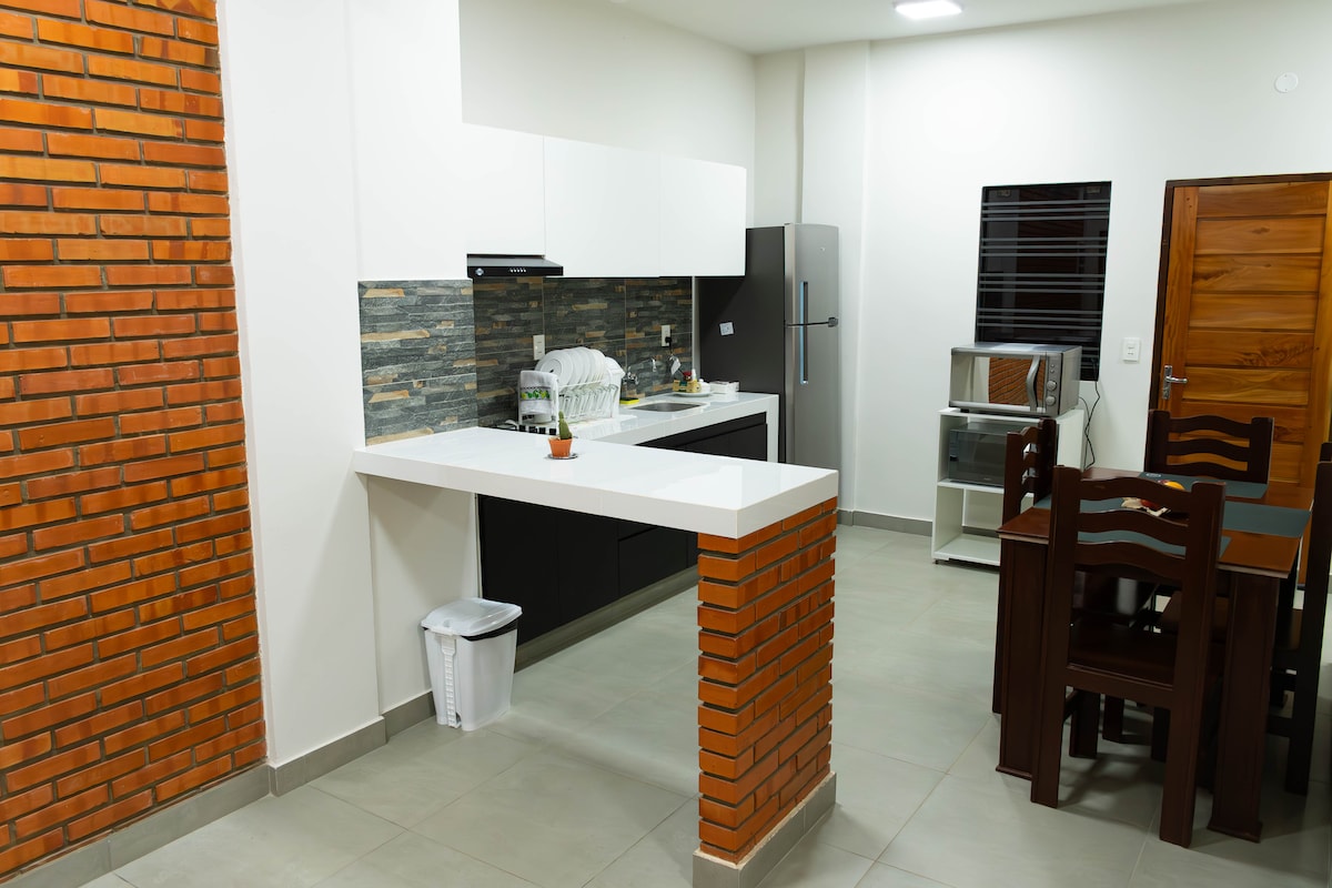 A modern kitchen setting is highlighted with a combination of white cabinetry and a textured brick accent wall. The kitchen is equipped with a stainless steel refrigerator, a microwave, and a dining area featuring a dark wooden table with matching chairs. Natural light enhances the spacious atmosphere.